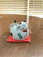 Front view of ceramic maneki-neko cup on red cushion showing overall shape, colors and decals