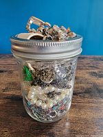 Front view of clear glass jar showing assorted jewelry pieces and metal band lid