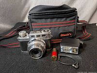 Front view of camera showing Schneider-Kreuznach Xenar 50 mm f/2.8 lens, flash unit, cable, red container and Black’s bag