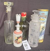 Group shot of all five items: two clear glass decanters with stoppers, one white floral glass decanter with stopper, one green glass bottle with cap, one Amarula tin