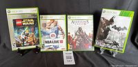 Front view of four Xbox 360 game cases displayed on stands: Lego Star Wars The Complete Saga, NBA Live 10, Assassin's Creed II, Batman Arkham City