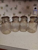 Four clear quart canning jars, three Ball Ideal and one Atlas E-Z Seal, showing embossing, lids, and wire bales