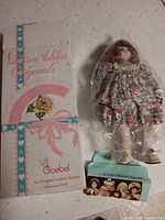 Porcelain doll sealed in plastic alongside original Victoria Ashlea Originals Goebel box and collector pamphlet