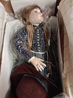 Doll inside box with plastic wrap showing face, blouse, vest and hat