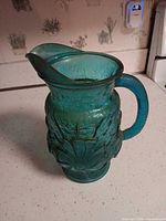 Full view of teal-blue glass pitcher showing floral relief, handle and spout