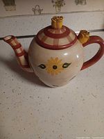 Side view of cream ceramic teapot with red handle, sunflower motifs, checkered spout, rosebud lid finial