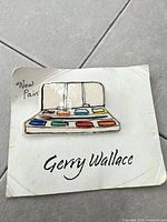 Front of ceramic brooch showing paint-palette design and artist name card