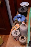 Photo showing seven assorted pottery crocks of various sizes on a wooden surface placed close together; includes a blue crock with lid and smaller cream crocks with stoppers and lids.