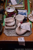 Photo showing assorted decor pieces including hand-painted floral cup and saucer, white ceramic creamer on rectangular base, white ceramic swan figurine, and blue and white porcelain box in the background.