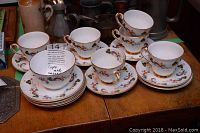 Photo of the full set showing six cups on saucers and five dessert plates arranged on a wooden surface with a sugar bowl and milk pitcher visible in the background.