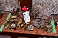 Overview of small brass and metal figures including animal figurines, bells, vases, and mineral specimens arranged on a wooden surface with lace curtains in the background.