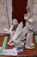 Photo showing all four Nao by Lladro figurines arranged with a lace curtain background. Front center is the boy figurine with large goose spreading wings.