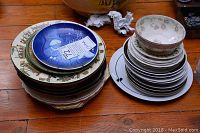Stack of assorted vintage decorative china plates on wooden floor, including blue and white plates and floral pattern bowl on top.