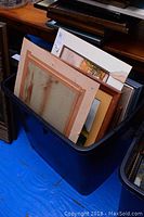 Bin filled with framed pieces of art in various sizes, showing edges and backs of frames and mats.