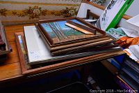 Stack of eight framed artworks in various sizes and styles sitting on a wooden surface, with visible frames and edges.