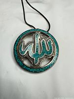 Front view of pendant showing turquoise mosaic calligraphy and border with attached leather cord