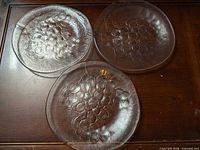 Three clear glass plates with grape motif on wooden surface
