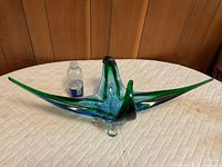 Tri-winged green and blue blown glass centerpiece beside bottle for scale