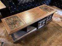 Top and front view of rectangular wood coffee table with glass-covered carved panels, central wood panel, carved cabinet doors, and open shelf