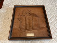 Front view of framed wood relief carving with outhouse design and title plate