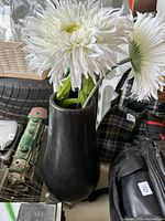 Black ceramic vase holding two artificial white flowers, surrounding objects for scale.