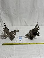 two silver metal rooster figurines with tape measure for scale