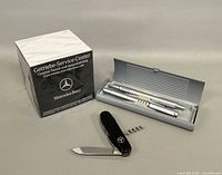 Overall lot showing sealed promotional box, open pen case with two writing instruments, and black pocket knife