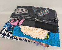 Stack of several scarves showing skull, pink motif, black fringe, chevron, turquoise leaf, beige pom-pom, paisley patterns