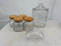 Group view of four small square jars with wood lids, large cylindrical jar with glass lid, and faceted covered dish