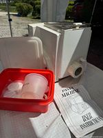 Side view of white electric pasta maker with accessory box, measuring cup, extrusion discs, instruction booklet visible