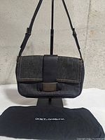Front view of denim pochette with leather trim and metal plate, dust bag below