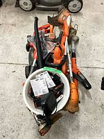Lot overview showing multiple orange yard tools, mower deck, bucket of batteries