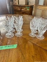Groups of stemmed cordial glasses and tumblers on wooden table; green tag noting 24 glasses