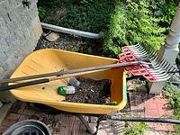 Yellow wheelbarrow with soil, two red-headed rakes, jar of pellets