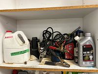 Shelf with white jug, tangled jumper cables, Toyota 0W-16 oil bottle, other fluid containers