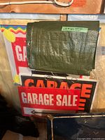 Folded green tarp resting on stack of Garage Sale yard signs with wire stakes visible