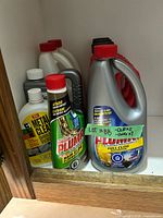 Group view of cleaners inside cabinet: Plumr jug, Plumr hair clog bottle, Metal Clear, calcium-lime jug and additional bottles