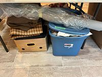 Overall view of cardboard box, blue Sterilite tote and plastic-wrapped fabric roll under table
