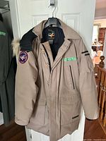 Full front view of tan Canada Goose parka on hanger showing pockets, buttons, hood and sleeve pocket