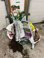 Stack of multiple soil bags against garage door, visible brands Voila, Golfgreen, Premier, generic bags, soil spilled on floor