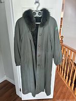 Front view of full-length olive green coat on hanger showing button closure, fur collar, pocket placement