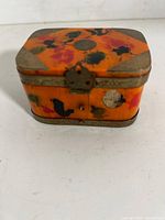 Front angled view of orange lacquer trinket box showing painted floral motifs and brass clasp