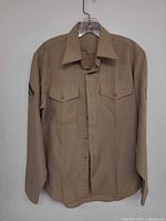 Front view of khaki military shirt on hanger showing pockets, buttons, left-sleeve chevron patch
