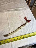 Full view of sword in scabbard beside ruler showing 16 in length, red tassel visible