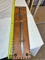 Full view of dagger mounted on leather plaque with measuring tape for scale