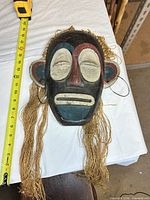 Front view of painted wooden mask showing colors, facial features, raffia fringe and measuring tape for scale