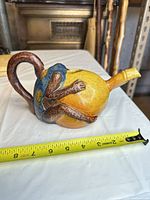 Side view of yellow ceramic teapot with brown monkey handle and blue vest detail, tape measure shows length about 6 in.