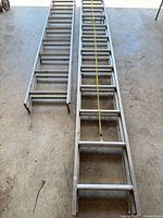 Two aluminum extension ladders side-by-side on floor showing rungs, rails, rope and foot caps