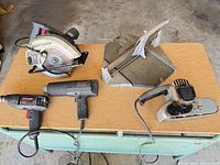 Group shot of five corded tools on table: circular saw, heat gun, drill, belt sander, saw base
