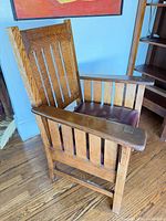 Front angled view of oak armchair showing slatted sides, leather seat, armrest inlay